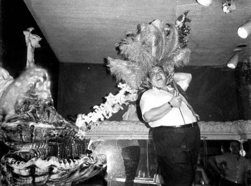 Vintage photo of Boston bar owner Phil Baione in his bar, decorated for the holidays. He is laughing and holding a large crown of ostrich feathers on his head like a hat.
